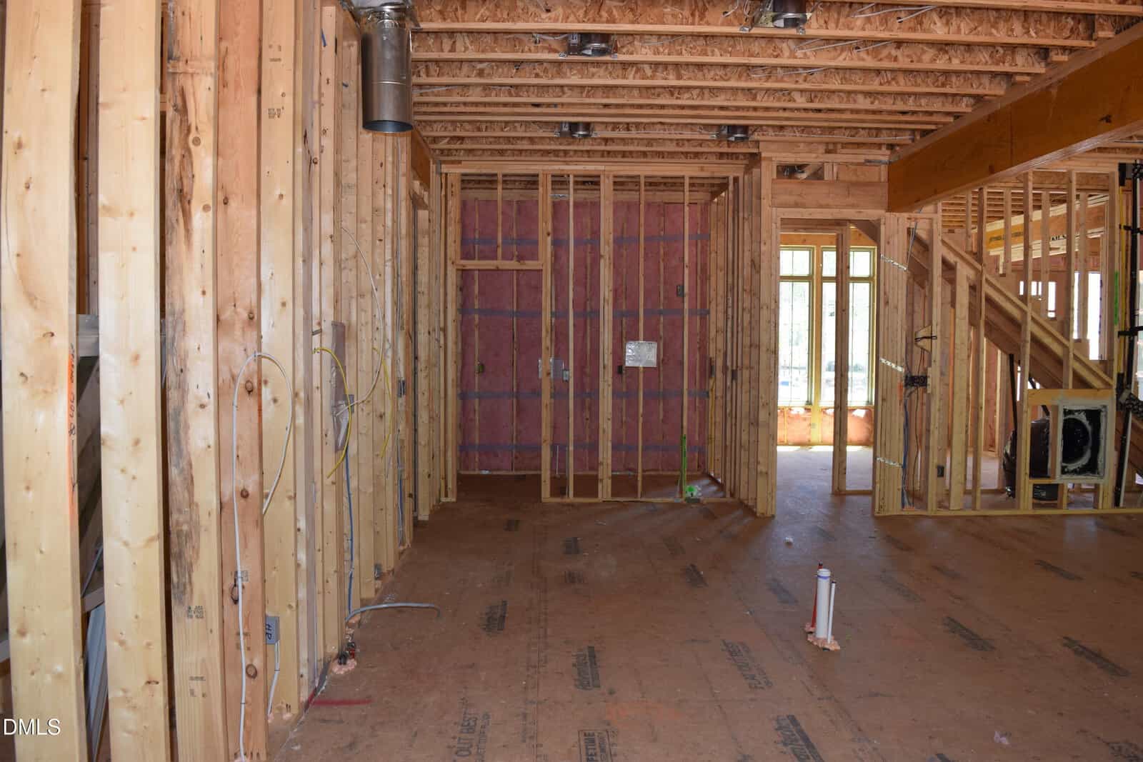 Interior of a house under construction showing exposed wooden framing, insulation in some walls, unfinished floors, and uninstalled electrical wiring. Natural light enters through open windows and doors.