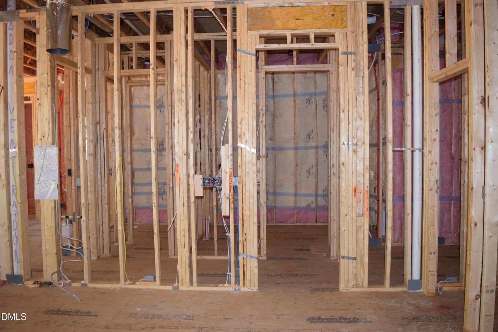 Wood framing of a house under construction, showing exposed studs, unfinished walls, and electrical wiring. Pink insulation is visible in some wall sections. No drywall or flooring has been installed yet.
