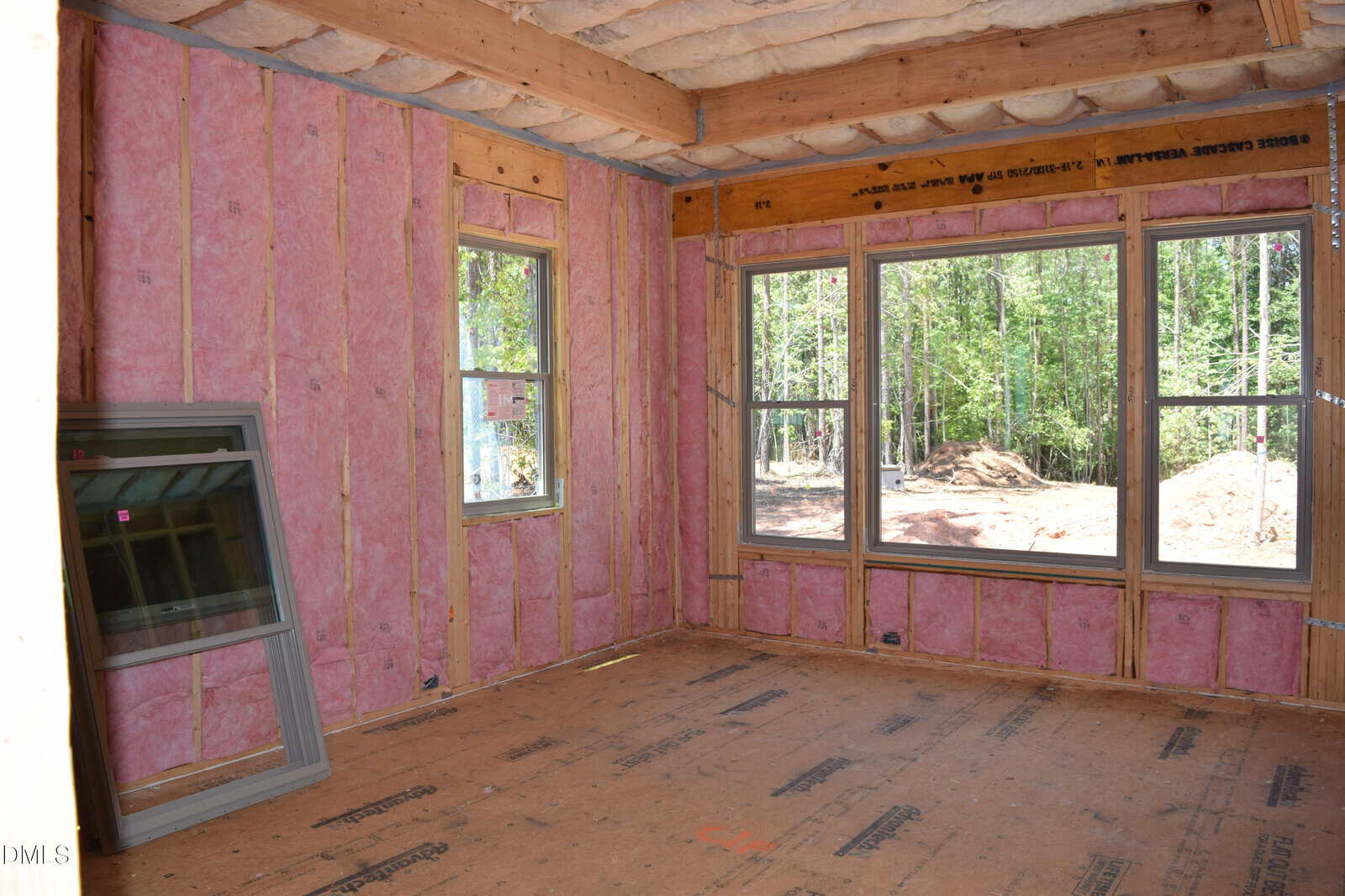 Room under construction with exposed pink insulation in the walls and ceiling, large windows letting in sunlight, and an extra window frame leaning against the wall. Trees and dirt are visible outside through the windows.