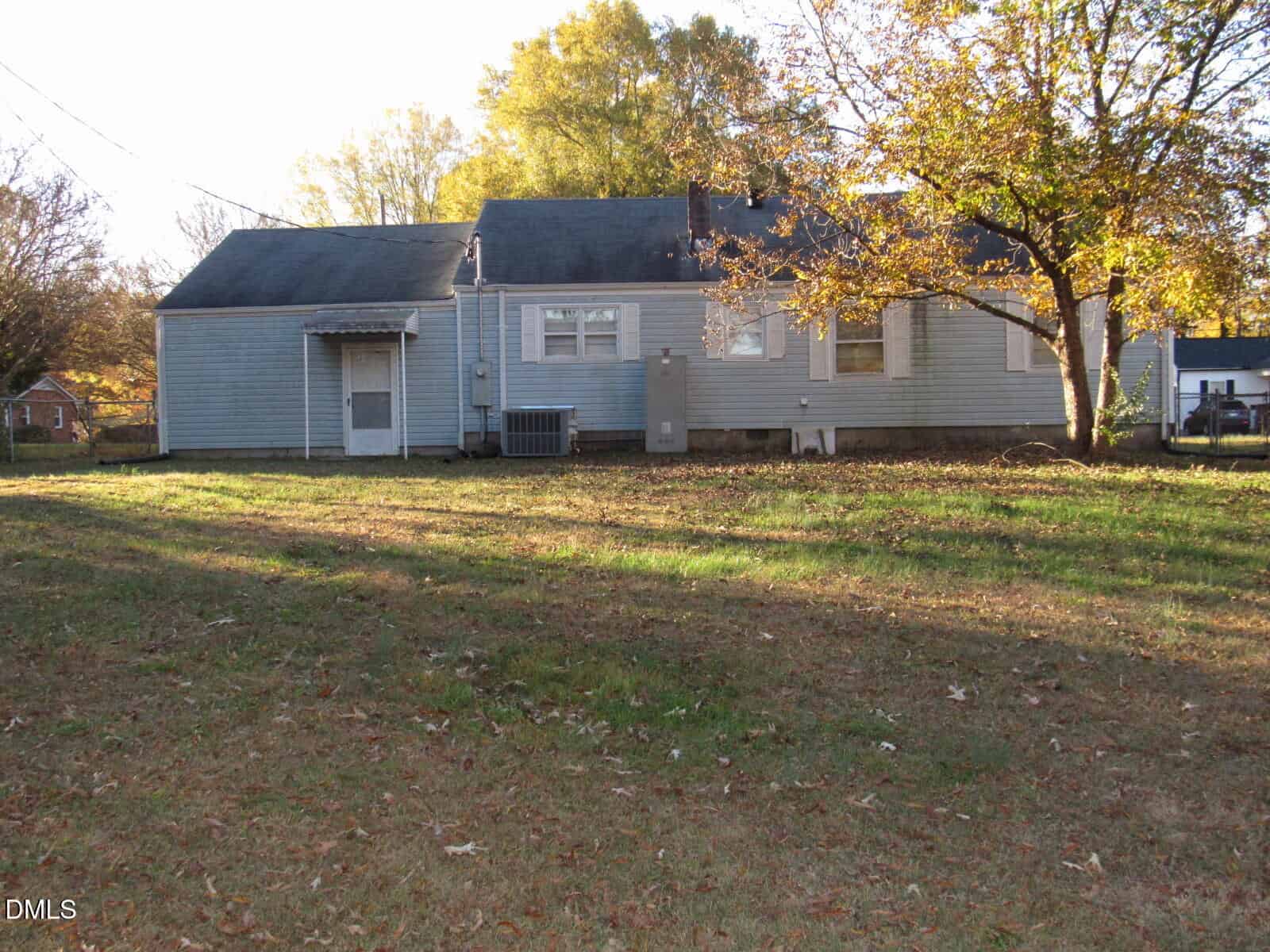 A single-story blue house with a gray roof sits in a grassy yard with scattered autumn leaves, bordered by trees with yellow and orange foliage.