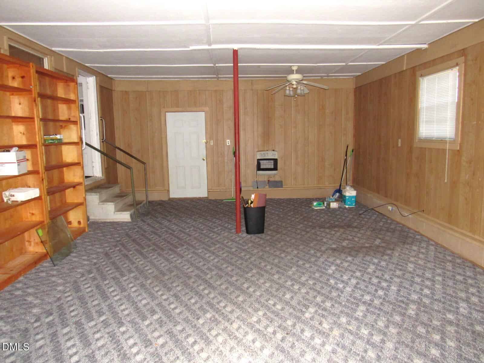 A basement room with wood-paneled walls, carpeted floor, a red support pole, built-in wooden shelves on the left, a ceiling fan, cleaning supplies in one corner, and a window on the right wall.