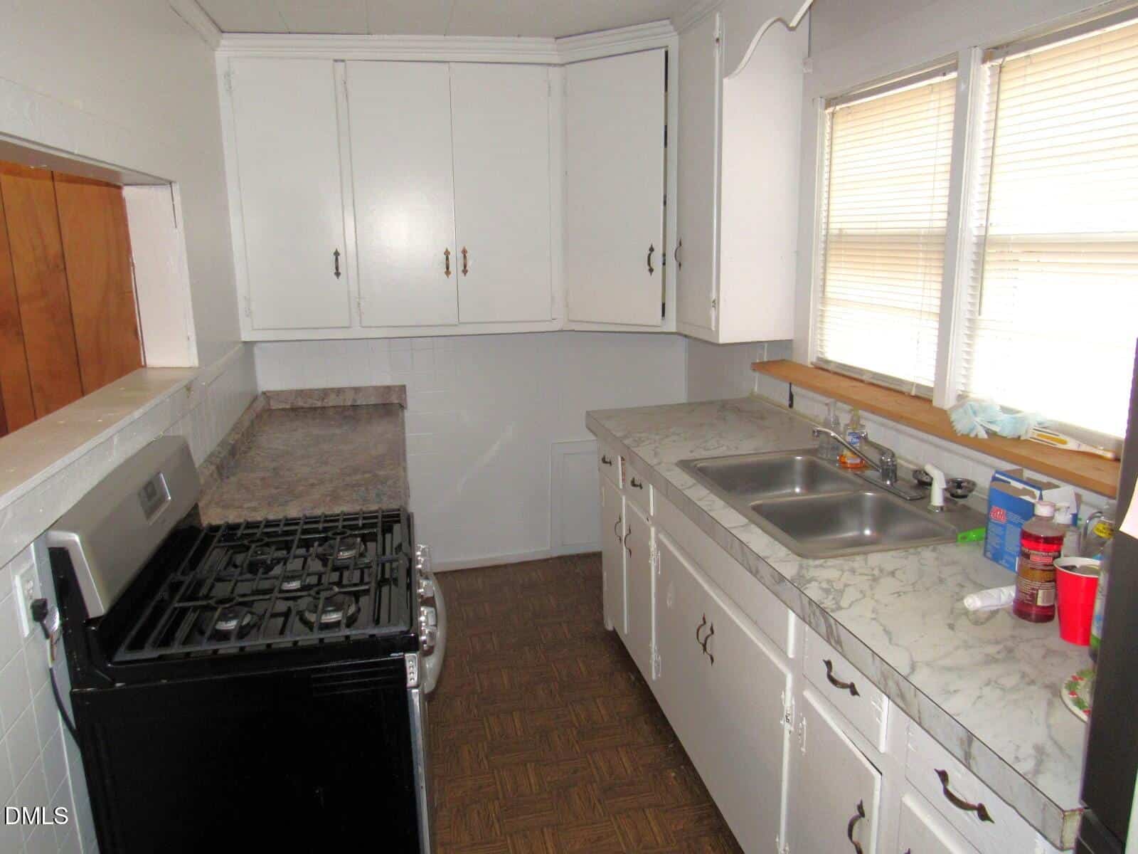 A small galley kitchen with white cabinets, marble-patterned countertops, a stainless steel gas stove, double sink, and two windows with blinds. Various cleaning supplies and items are on the countertop near the sink.