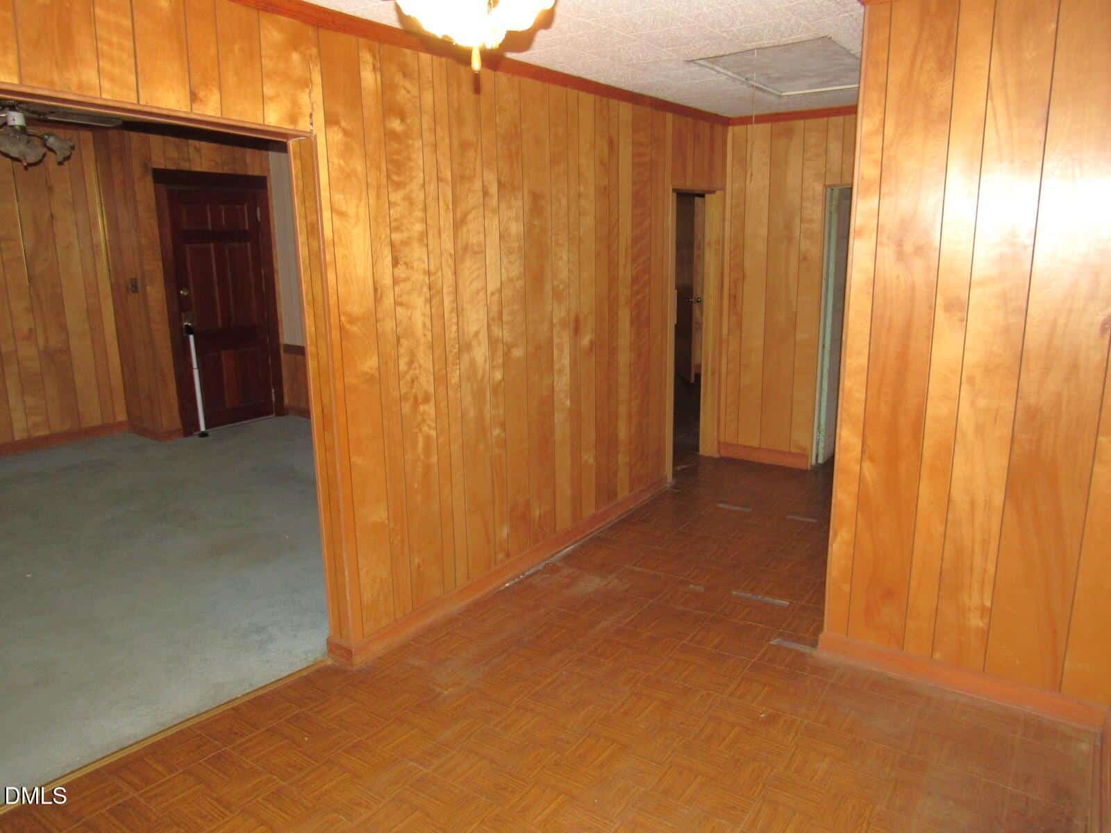 A room with wood-paneled walls, parquet flooring, and an adjacent carpeted room visible through an open doorway. There is a closed brown door in the background and a ceiling light fixture overhead.