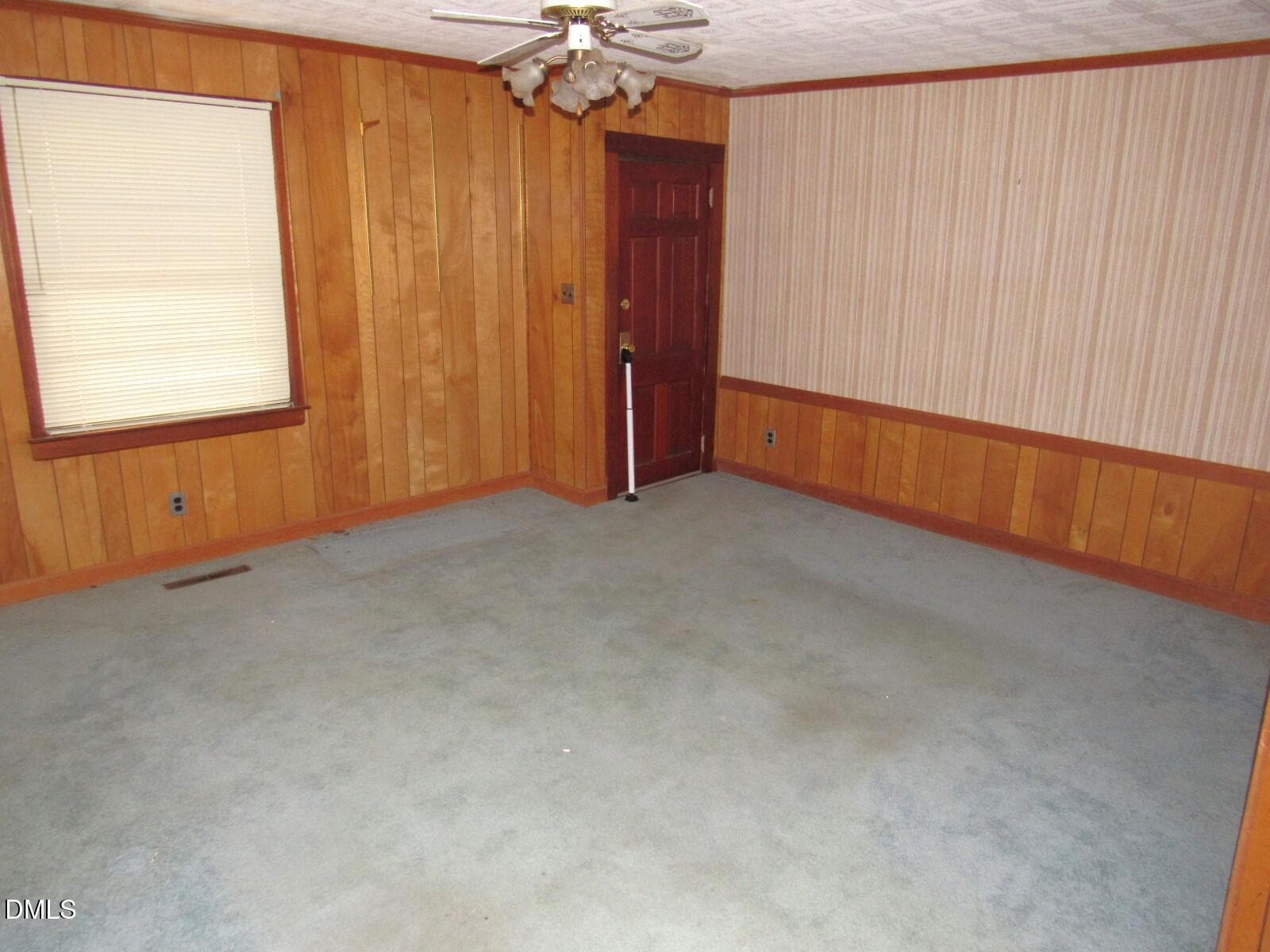 Empty room with wood-paneled walls, a single window with closed blinds, a red wooden door, blue carpet flooring with visible stains, and a ceiling fan with light fixture. A white cane rests against the wall near the door.