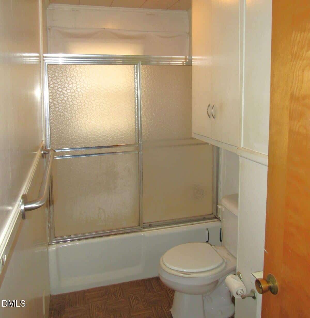 A small bathroom with a bathtub and sliding glass shower doors, a white toilet, white cabinets above the toilet, a towel bar on the left wall, and a wooden door partially opened.