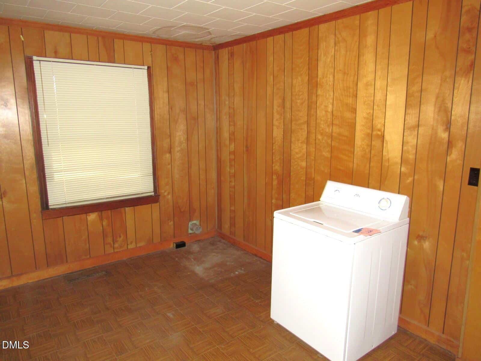 A small room with wood panel walls, a single window with closed blinds, and a white washing machine in the corner on a patterned linoleum floor. The room appears mostly empty and has some discoloration near the baseboard.