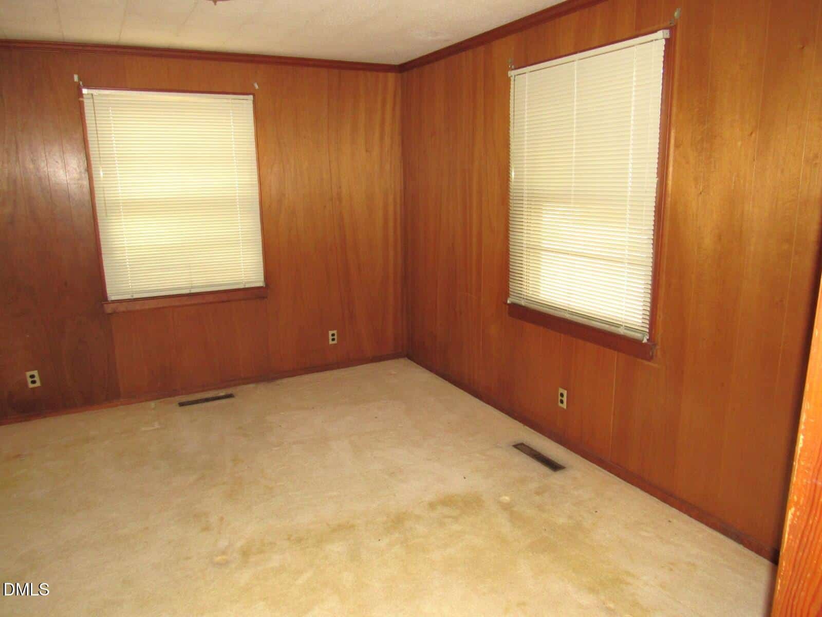 An empty room with wood-paneled walls, two windows with closed blinds, beige carpet, and visible floor vents. Electrical outlets are present on the walls. The room appears clean and undecorated.