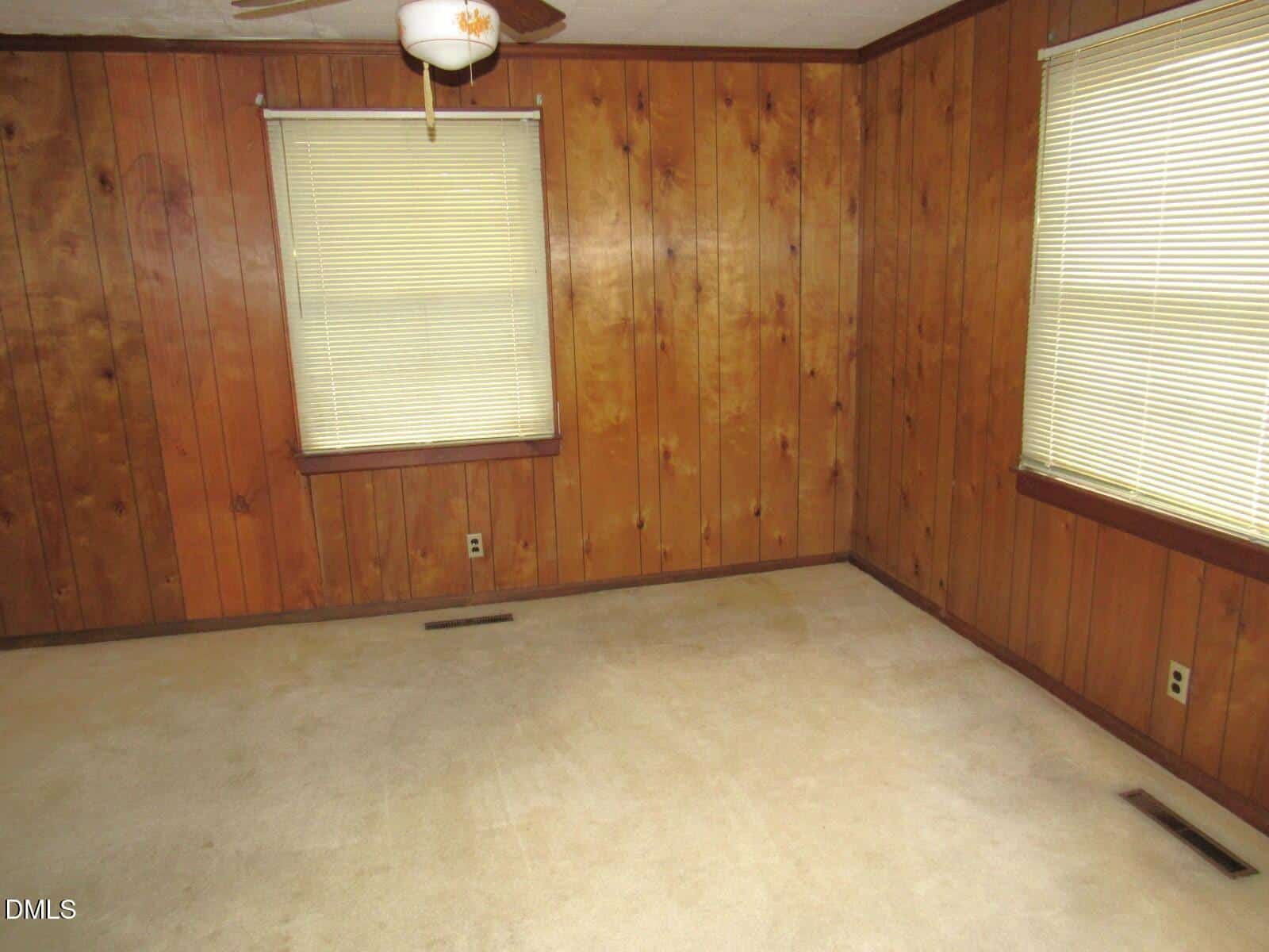 Empty room with wood-paneled walls, beige carpet, two windows with closed blinds, a ceiling fan with a light, and visible electrical outlets and vents.
