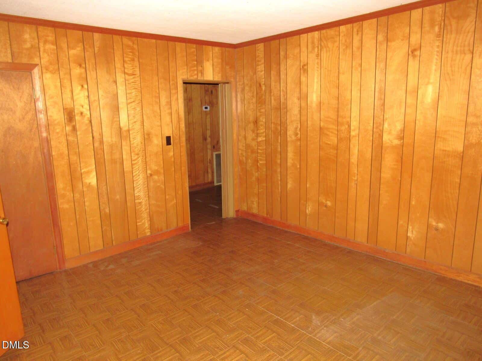 Room with wood-paneled walls, parquet-style wood flooring, and a white ceiling. There is one open door leading to another room and one closed door on the left side. The room appears empty.