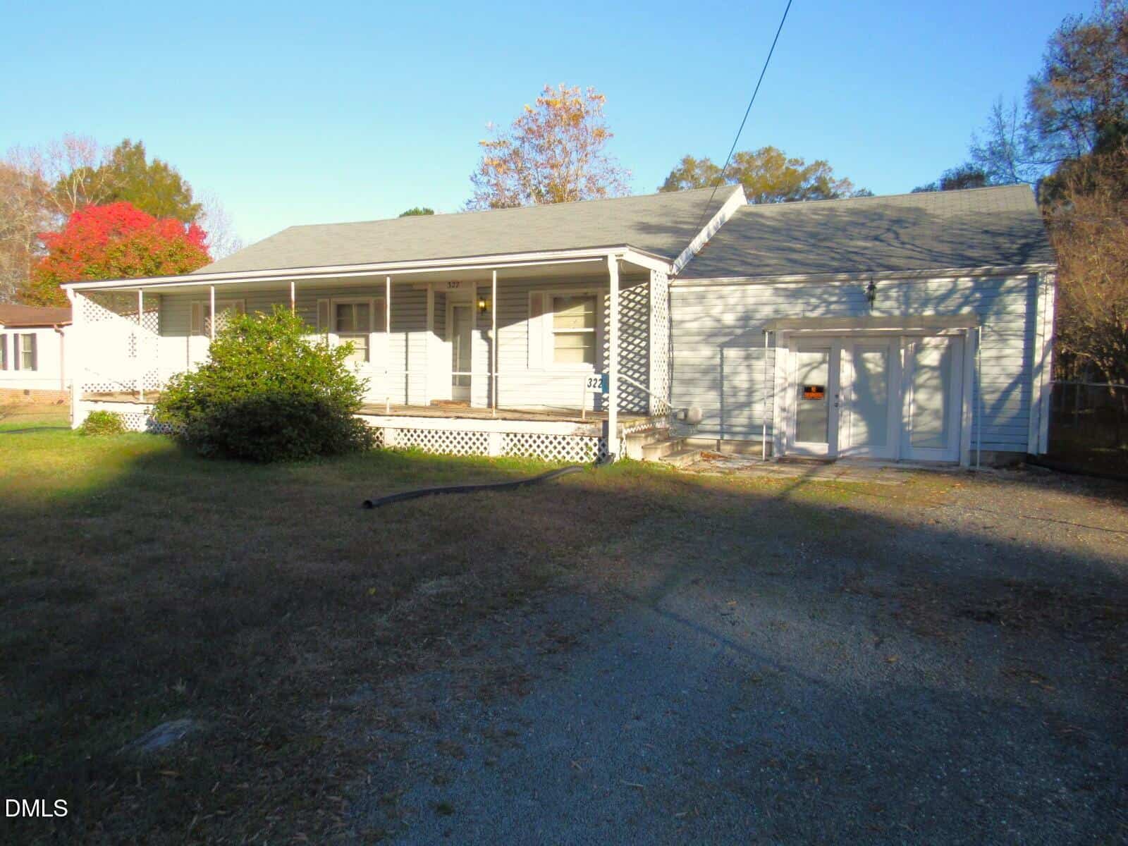Located at 322 Aiken Avenue in Creedmoor, this single-story house features a covered front porch, white siding, and an attached garage. A gravel driveway leads to the home, bordered by a dry lawn and trees under a clear sky.