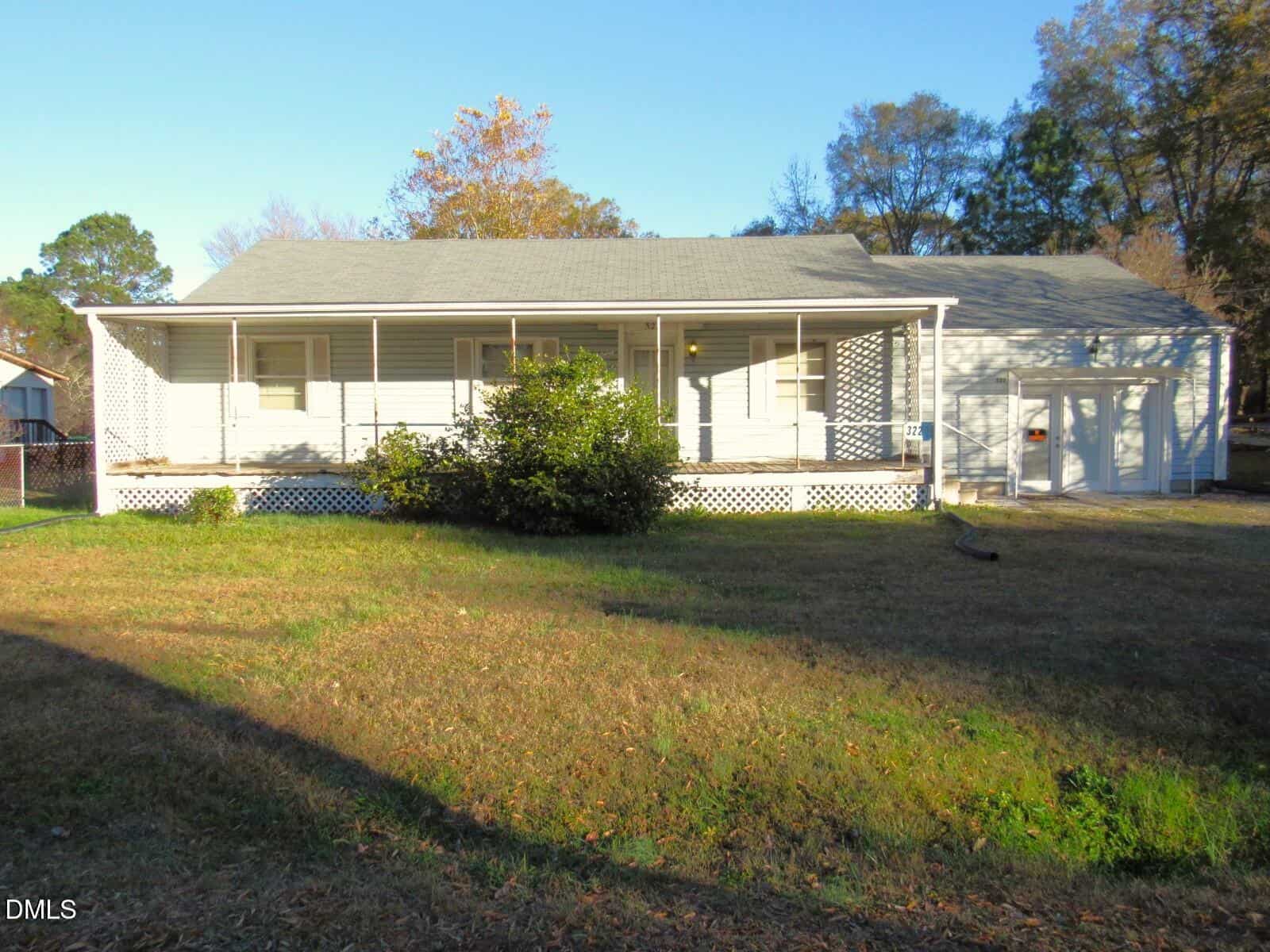 A single-story white house with a large covered front porch, lattice skirting, and a gray shingle roof, set on a grassy yard with some bushes and trees in the background.