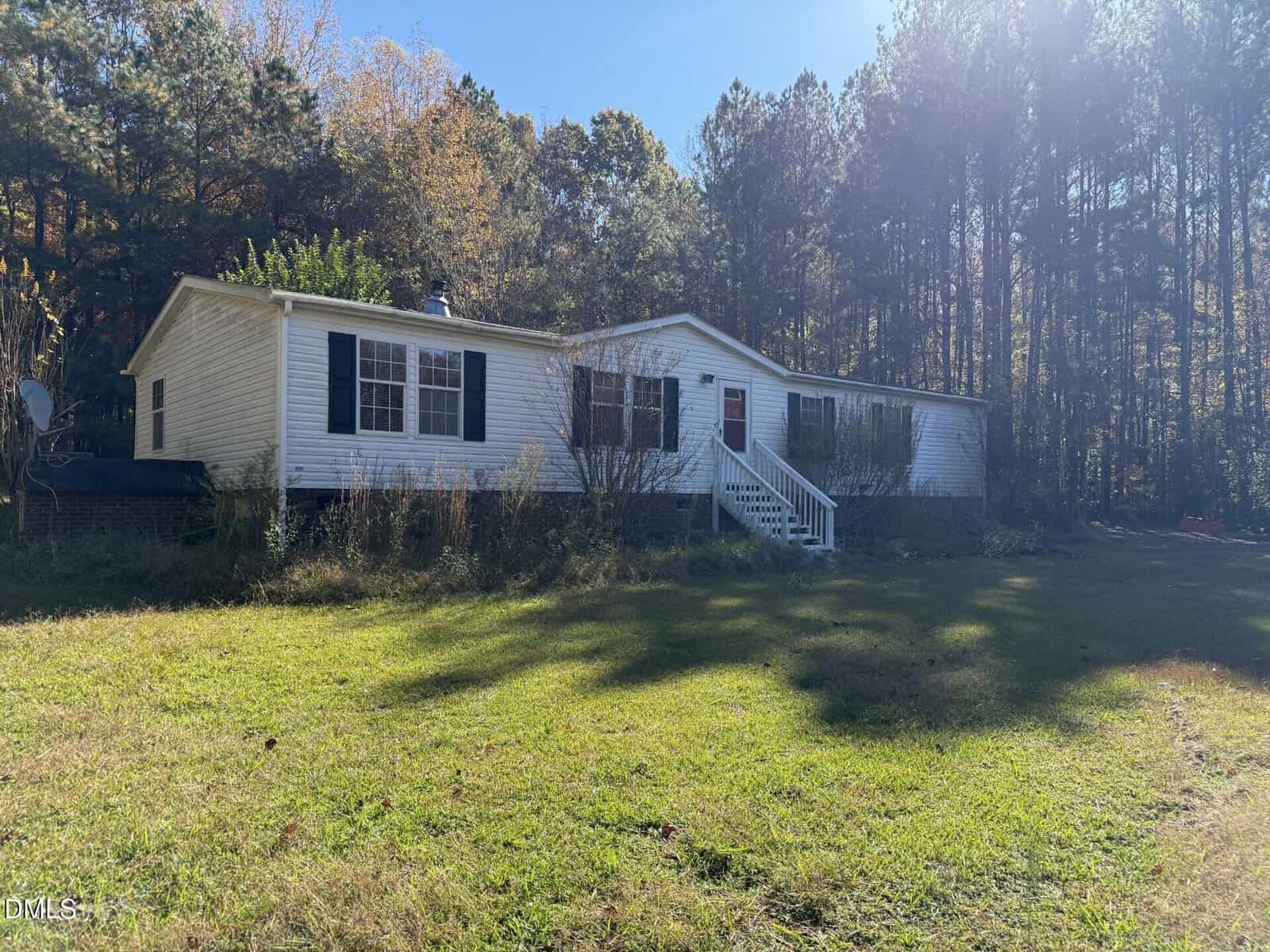 A single-story white manufactured home with black shutters sits in front of a wooded area. The house has steps leading to a red front door and is surrounded by grass and some overgrown plants. The sky is clear and sunny.