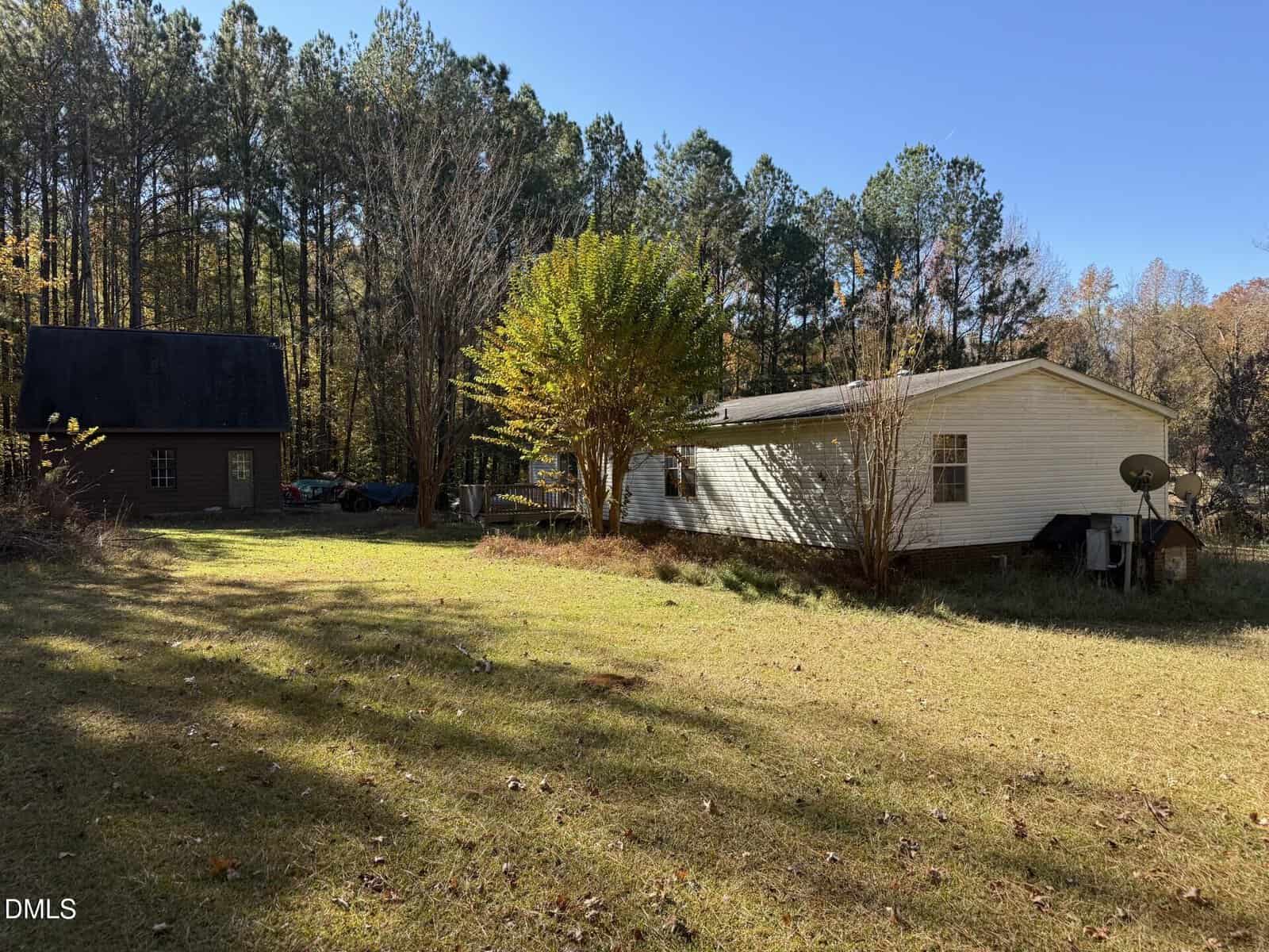 A white single-story house and a dark two-story building sit on a grassy lot bordered by tall trees under a clear blue sky. A satellite dish is attached to the house, and a tree with autumn foliage is in the yard.
