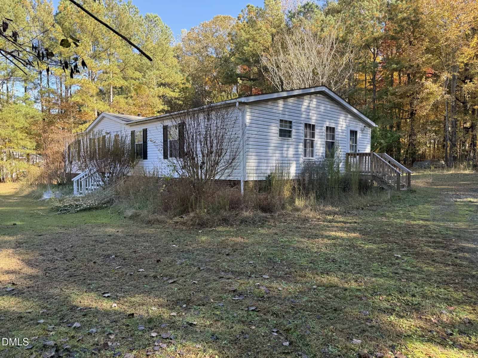 A white manufactured home with brown steps in a grassy yard, bordered by overgrown shrubs and surrounded by trees with autumn foliage under a clear blue sky.