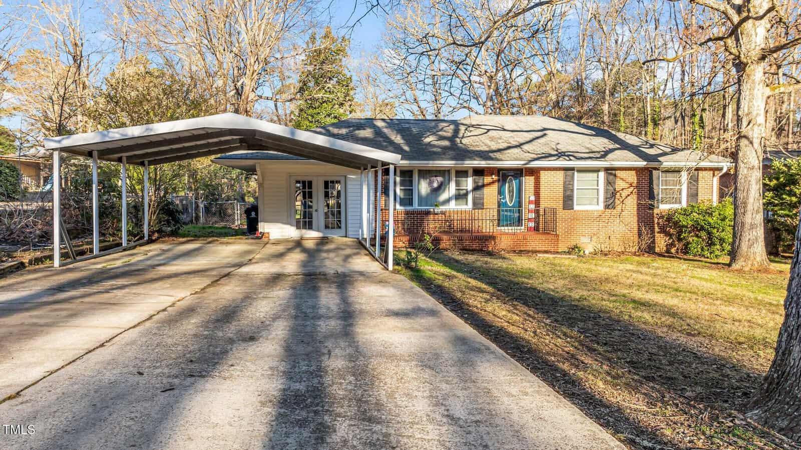 Single-story brick house with a blue front door, white trim, and attached carport. Concrete driveway extends from the street to the carport. Yard has grass, trees, and some shrubs near the house.
