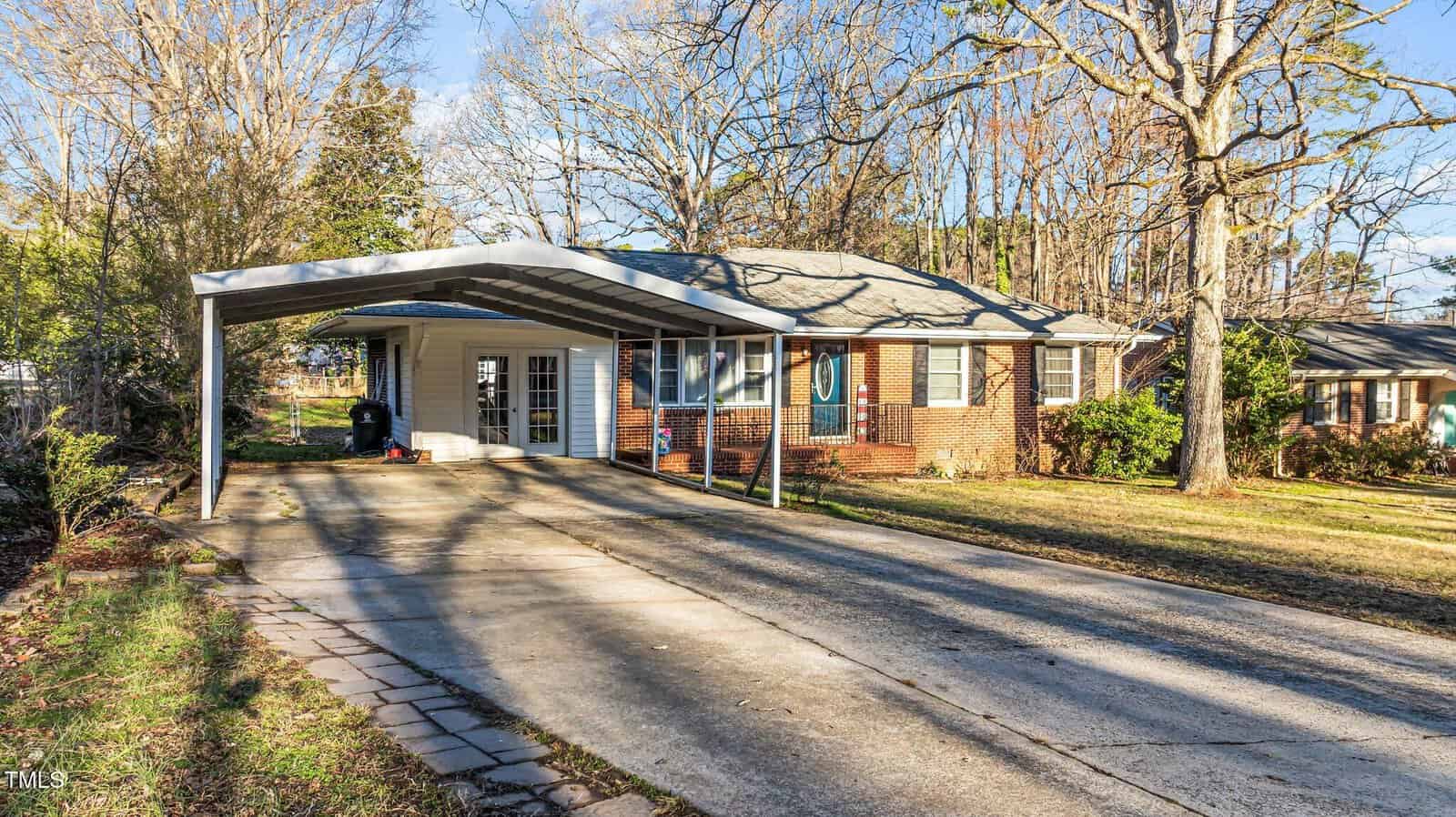 Single-story brick house with a gray shingle roof, large carport, and long driveway. Leafless trees and some green shrubs surround the building, and there is a lawn area in front of the house.