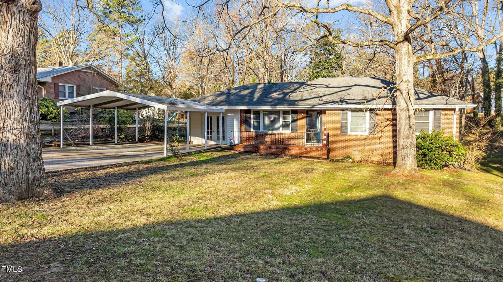 Single-story brick house with a gray shingle roof, covered carport on the left, and a grassy front yard with large trees. The home is surrounded by other houses and tall trees in the background.
