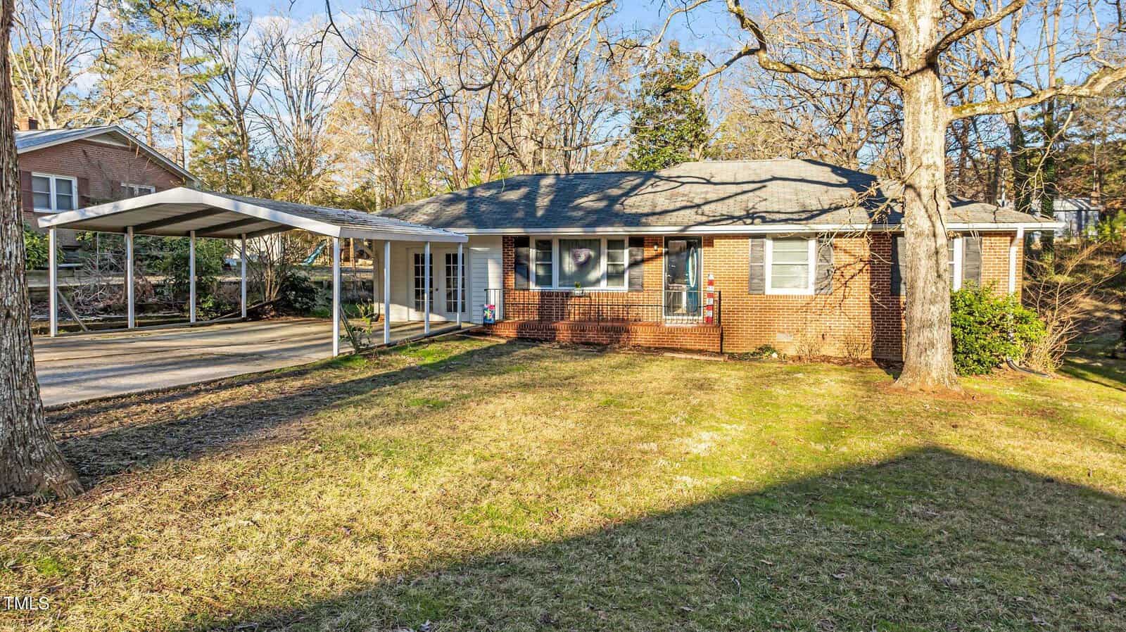 A single-story brick house with a gray shingle roof, large front windows, and an attached carport. The front yard is grassy with some patches of bare ground and bordered by mature trees.