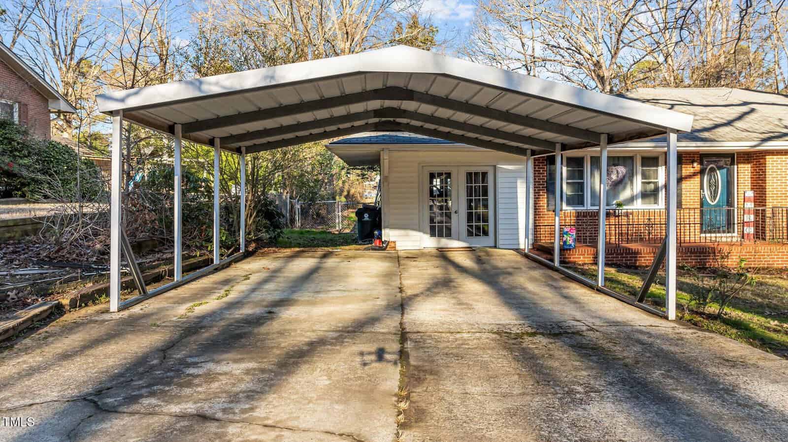 A wide, metal carport with a peaked roof stands over a concrete driveway next to a brick and siding house with French doors, on a clear day with bare trees in the background.