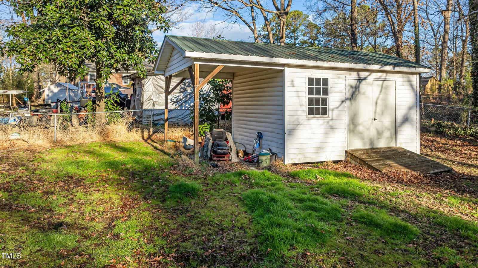 A white shed with a green metal roof stands in a fenced backyard. A ramp leads to the closed door, and a window is on one side. Lawn equipment and a lawnmower are stored under the small covered area beside the shed.