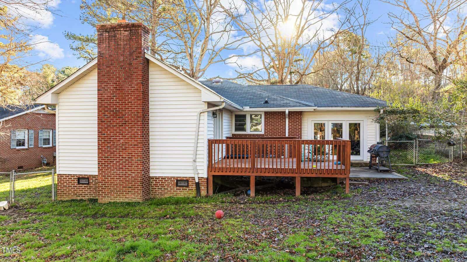 A single-story house with white siding and a brick chimney, featuring a wooden deck. The yard has patchy grass and a red ball, with leafless trees and another house visible in the background.