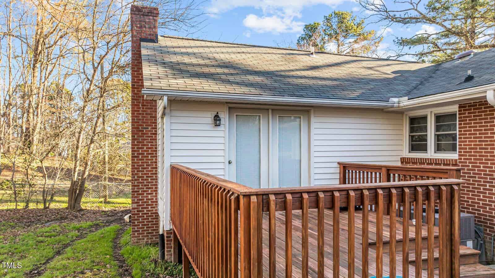 A small wooden deck with railing is attached to a house with both brick and white siding. French doors lead to the deck, and a chimney is visible on the left side. Trees and a grassy yard are in the background.