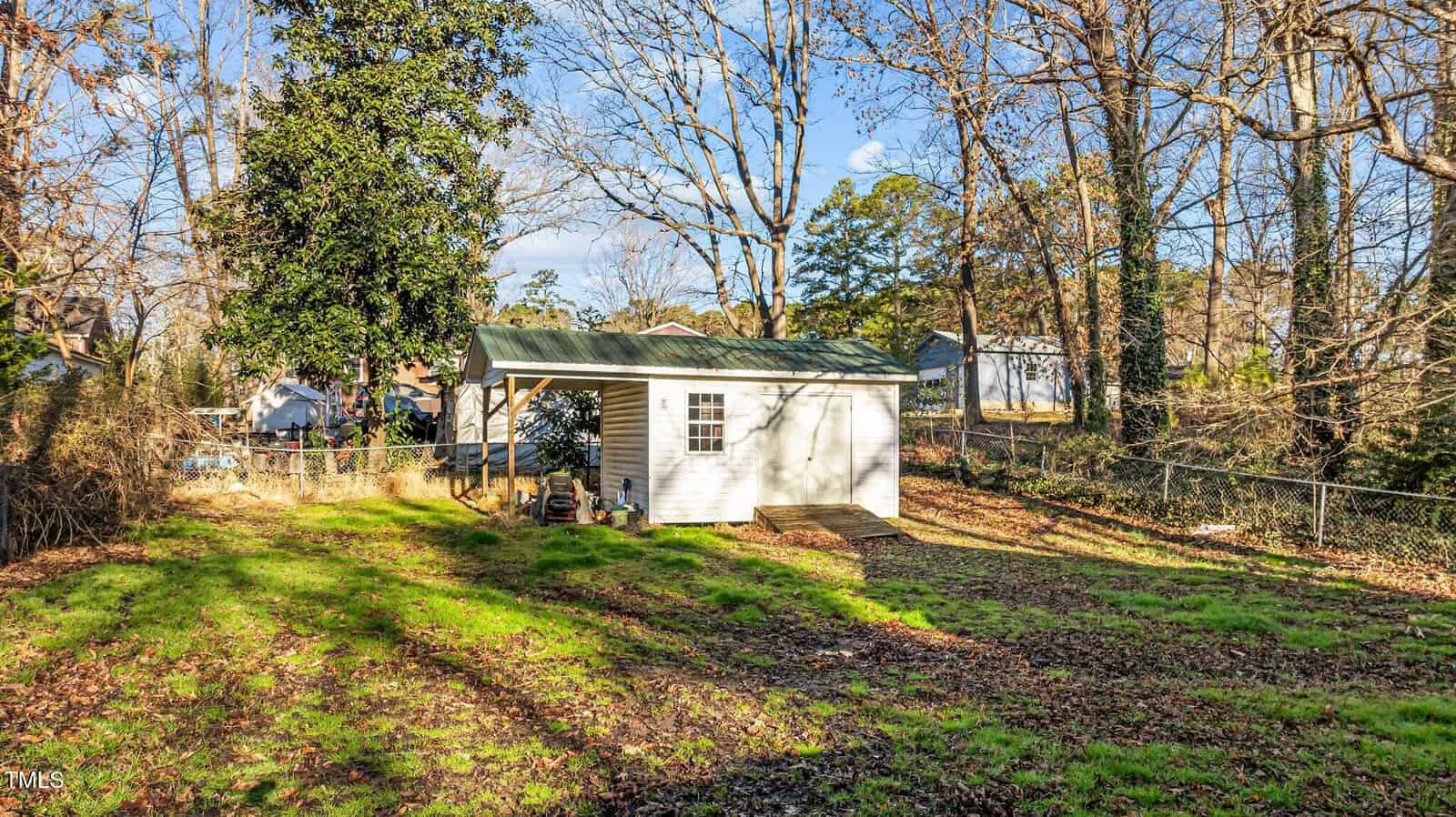 A white shed with a green metal roof sits in a fenced backyard. The shed has a small covered porch area, surrounded by trees with no leaves and a patchy grassy lawn. The weather is sunny with blue sky.