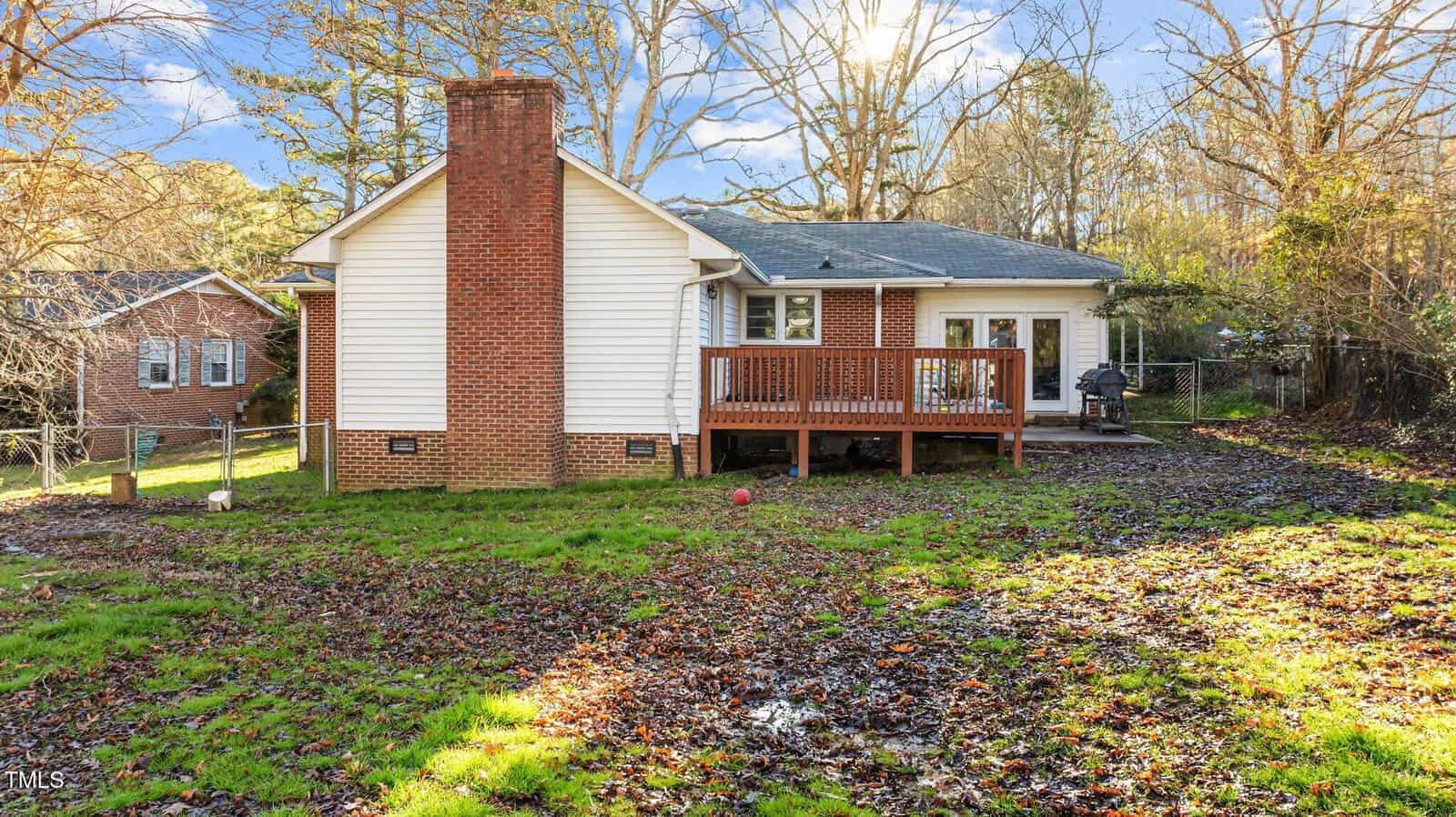A small house with white siding and a brick chimney, a wooden deck, and a fenced backyard with patchy grass and scattered leaves. Trees and another house are in the background.