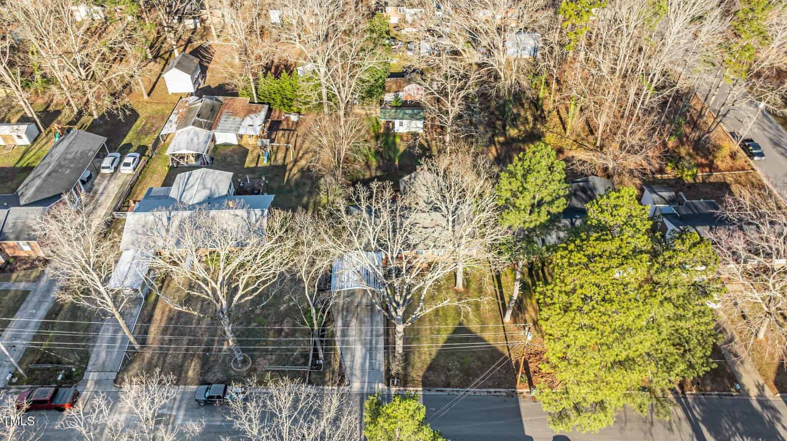 Aerial view of a suburban neighborhood with houses, driveways, leafless trees, parked cars, and intersecting streets on a sunny day.