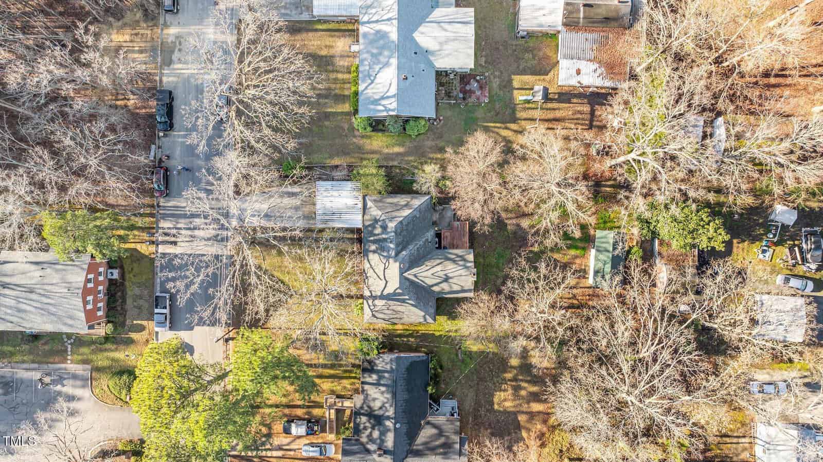 Aerial view of a residential neighborhood with houses, trees, cars, and lawns. Leafless trees cast long shadows, suggesting late autumn or winter season. Streets and driveways connect the homes.