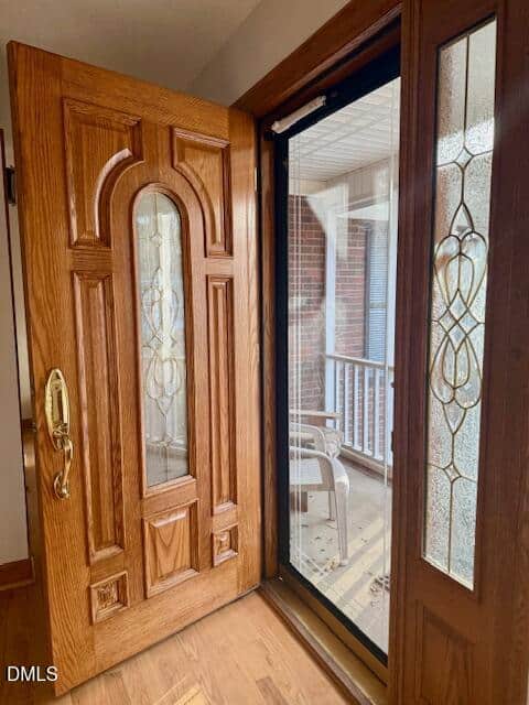 A wooden front door with decorative glass panels is open, revealing a glass storm door and a porch with white railings and a plastic chair outside. Sunlight brightens the entryway.