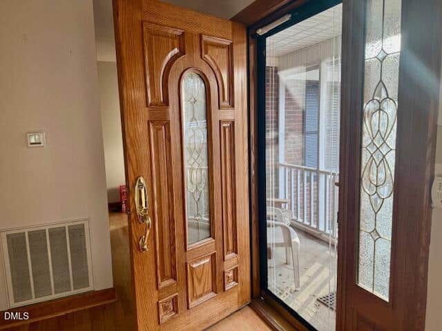 A wooden front door with decorative glass panels is open, revealing a glass storm door and a covered porch with chairs outside. Sunlight streams in, illuminating the entryway.