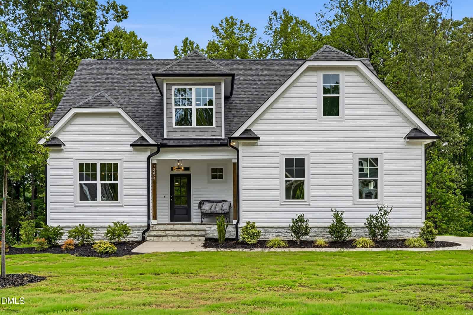 A modern white two-story house with a dark roof, multiple windows, a front porch, and neatly landscaped yard with green grass and shrubs, surrounded by trees.