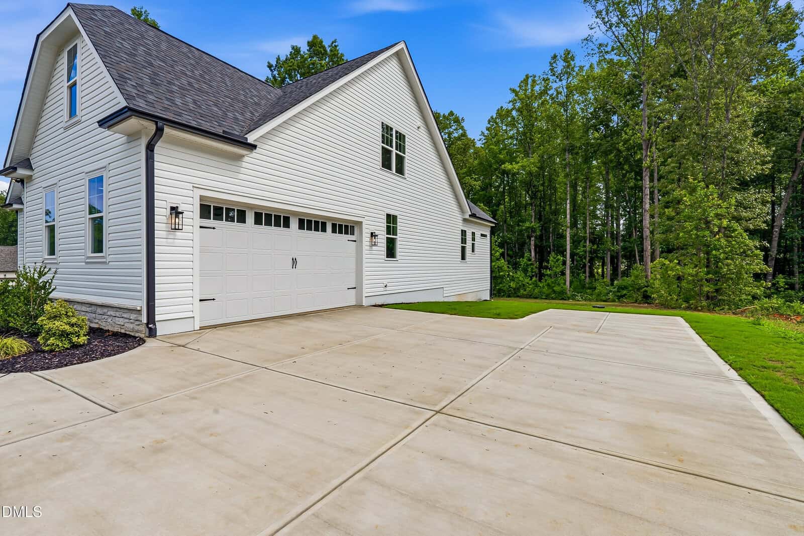 A modern white house with a large two-car garage and a wide, clean concrete driveway, surrounded by green grass and trees under a blue sky.
