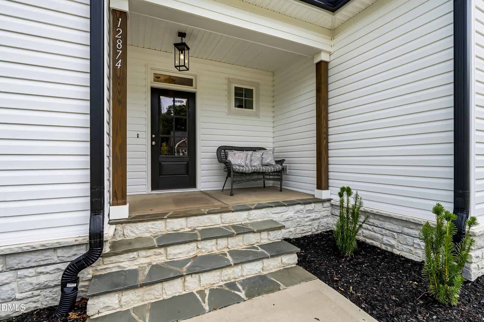 Front porch with stone steps, a black door, white siding, a black lantern, a wicker bench with cushions, and small green plants along the edge.