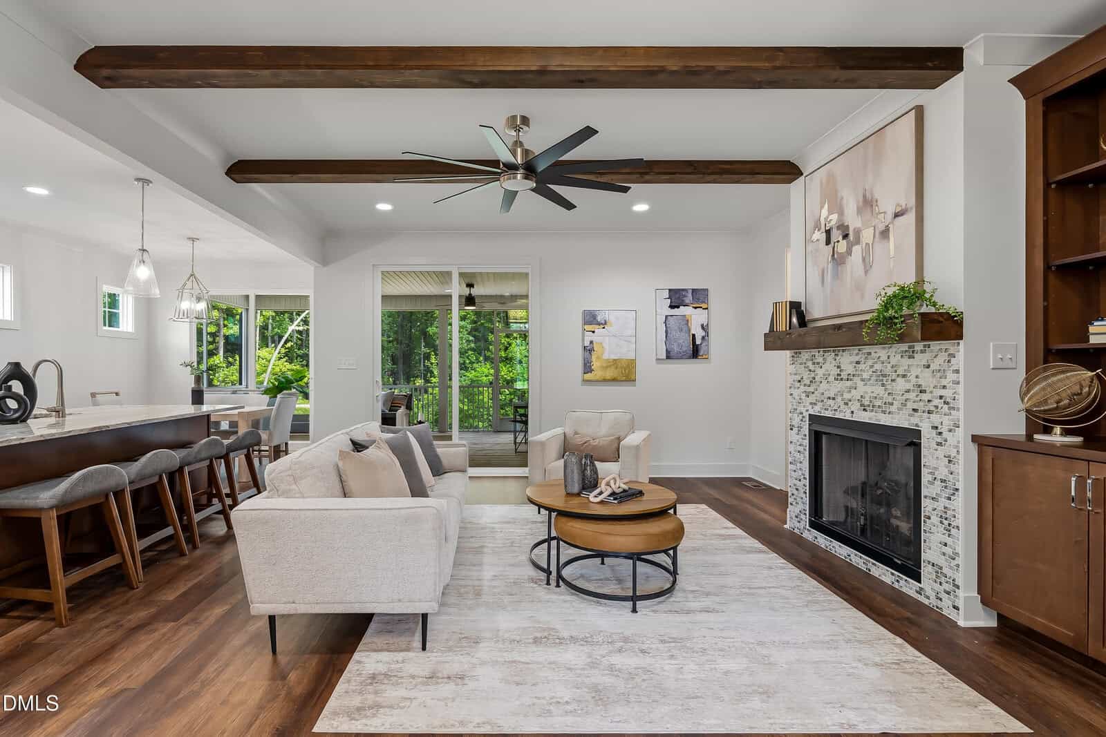 Modern living room with wood beams, neutral sofas, round coffee tables, a tiled fireplace, wall art, and large windows overlooking greenery. Open kitchen with barstools visible on the left. Wood floors throughout.