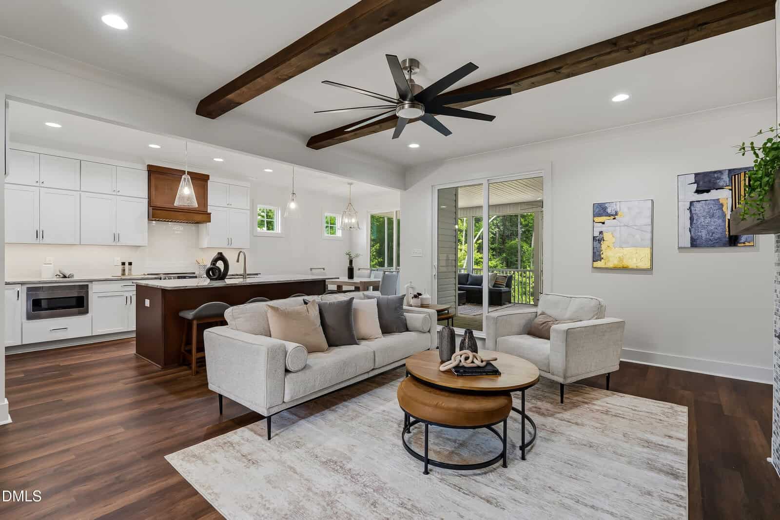 Modern open-concept living room and kitchen with exposed beams, neutral furniture, wood flooring, large ceiling fan, and sliding doors leading to a covered outdoor area. Contemporary decor and natural light fill the space.