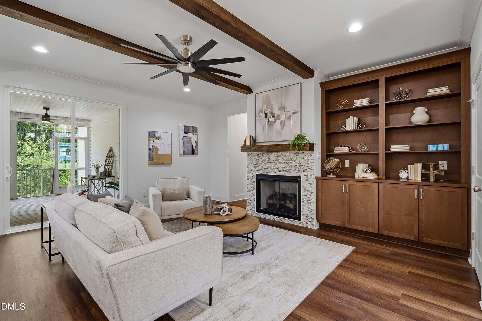 A modern living room with a beige sofa, armchair, fireplace, wooden shelves, ceiling fan, and exposed beams. Large windows and a door lead to a bright sunroom with greenery visible outside.