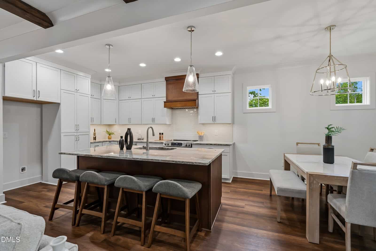 Modern kitchen with white cabinets, stainless steel appliances, a marble island with four gray barstools, pendant lights, a dining table with chairs, wood flooring, and small windows letting in daylight.