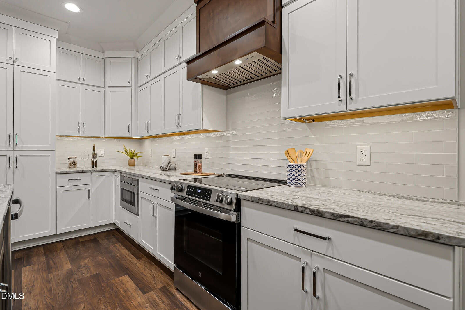 Modern kitchen with white cabinets, stainless steel appliances, marble countertops, wood flooring, and a plant on the counter. A stove is centered beneath a wooden range hood, with utensils in a holder nearby.