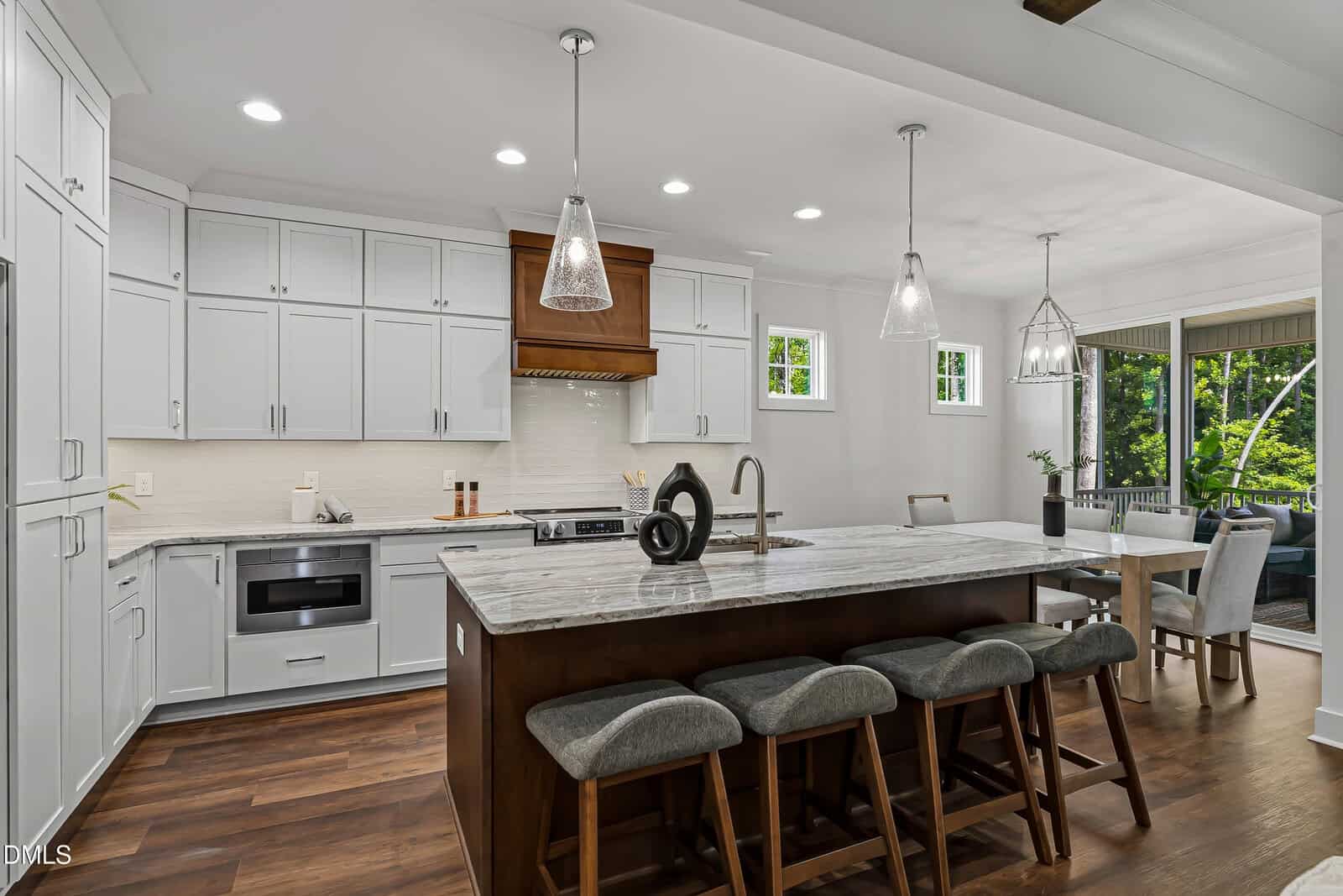 Modern kitchen with white cabinets, marble island with four gray stools, stainless steel appliances, wood floor, pendant lights, and adjacent dining area with large windows overlooking greenery.