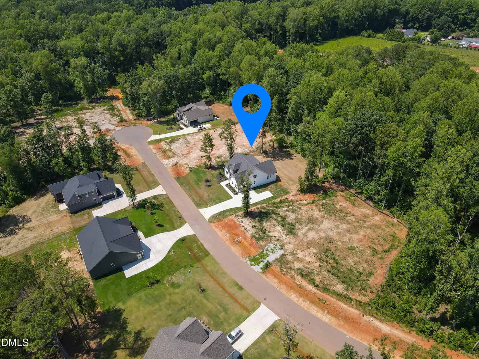 Aerial view of a suburban neighborhood with several houses, lawns, and surrounding trees. A large blue location pin is placed above the roof of a central house.