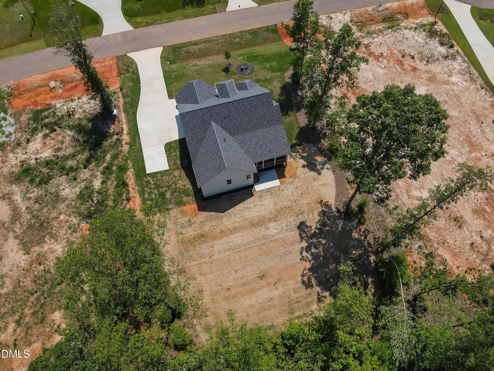 Aerial view of a newly built house with a dark roof, surrounded by bare earth and patches of grass, trees nearby, and driveways leading to the street.