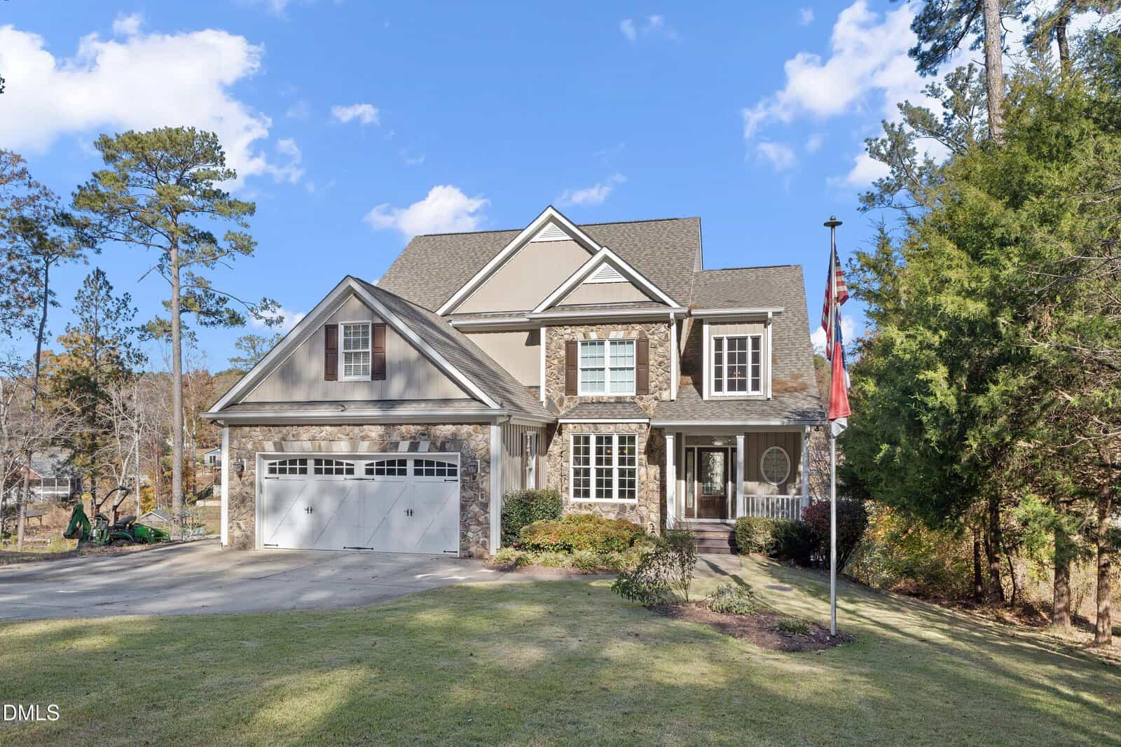 Two-story suburban house with a stone and beige exterior, a two-car garage, a front porch, manicured lawn, and an American flag on a pole. Trees and blue sky are visible in the background.