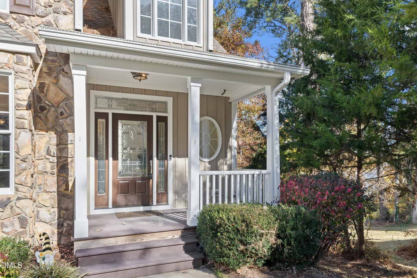 Front porch of a house with stone and beige siding, double glass front doors, white trim, a small covered area with a railing, stair steps, green shrubs, and a large tree to the right.