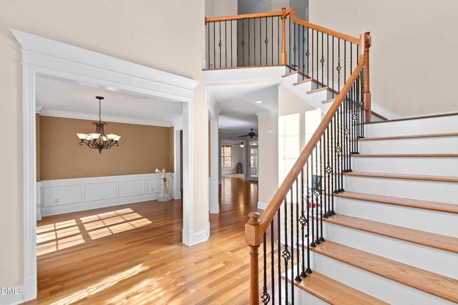 Bright foyer with hardwood floors, a wooden staircase with black iron railings, and an open dining room with wainscoting and a chandelier. Natural light fills the space, highlighting the warm, neutral walls.
