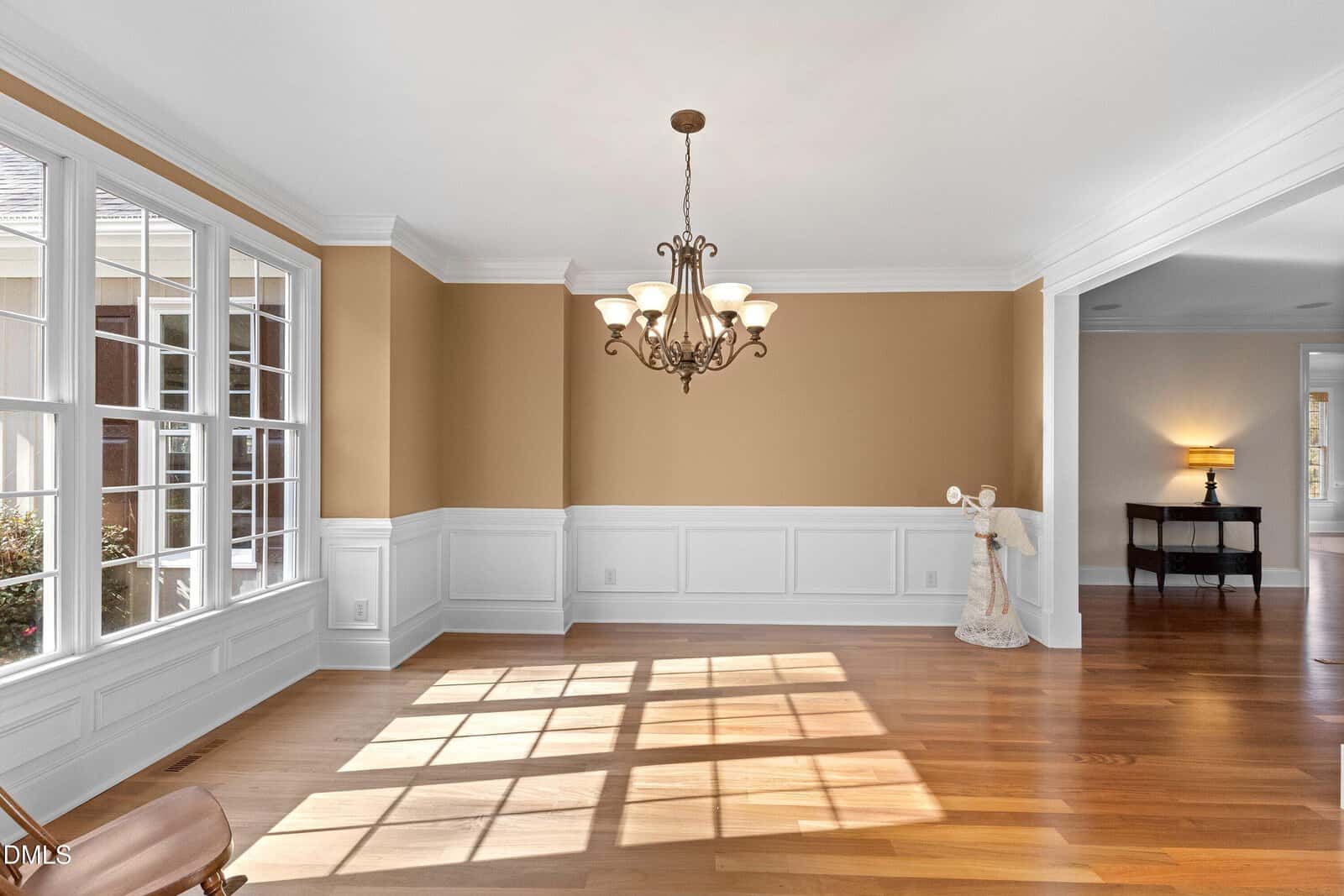 A bright, empty dining room with large windows, tan upper walls, white wainscoting, hardwood floors, and a decorative chandelier hanging from the ceiling. Sunlight streams in, casting shadows on the floor.