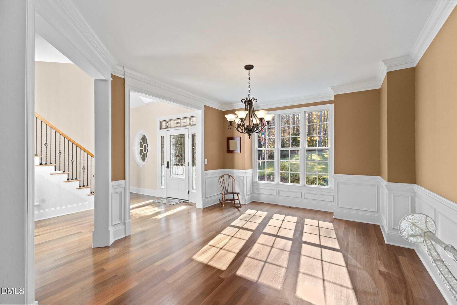Bright dining room with large windows, tan walls, white wainscoting, hardwood floors, and a chandelier. Sunlight streams through the windows, casting shadows on the floor. An empty chair sits by the window.