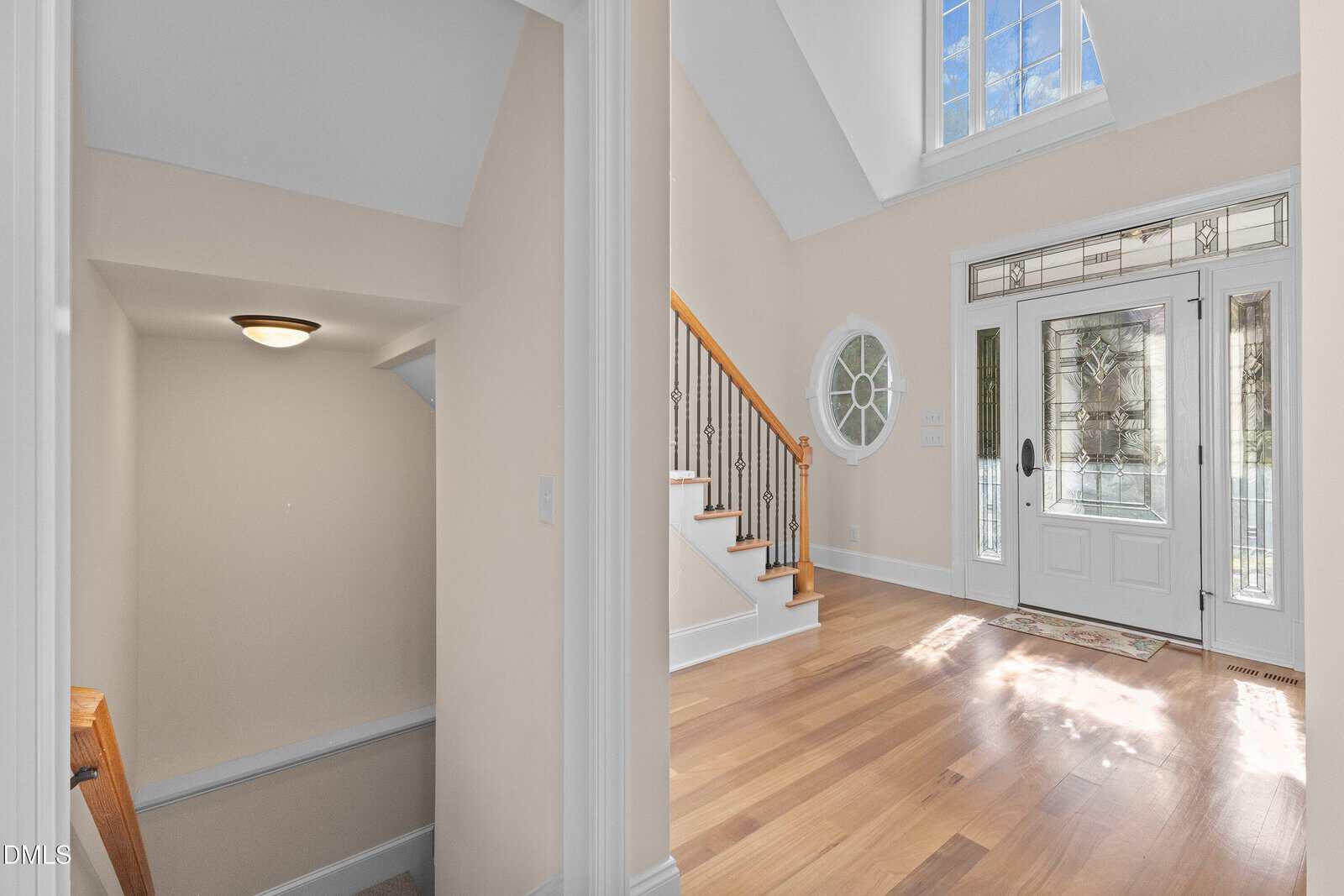 A bright foyer with hardwood floors, a large glass-paneled front door with sidelights, a decorative round window, and a staircase with a wooden handrail and black spindles. A stairway with a ceiling light is visible to the left.