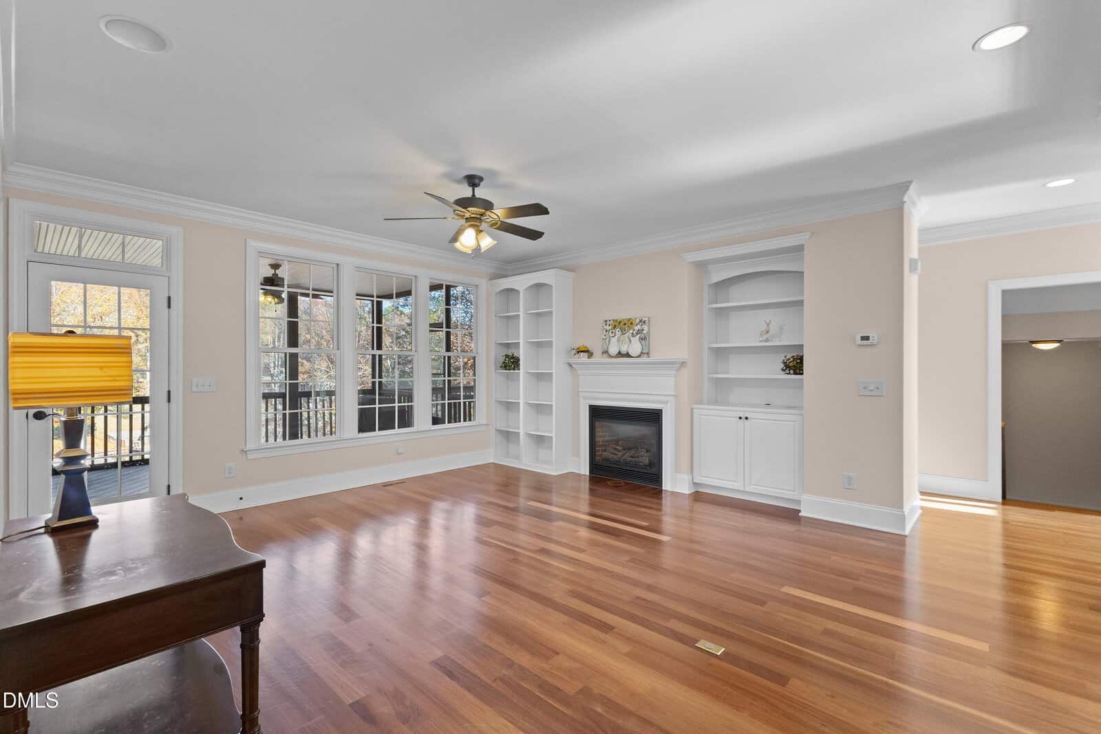 A bright, empty living room with hardwood floors, a ceiling fan, built-in white shelves, a fireplace, and large windows letting in natural light. A wooden desk sits to the side near a door leading outside.